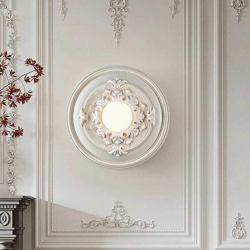 White LUXELY round wall light fixture with baroque-style detailing, adding elegance to a richly decorated wall.