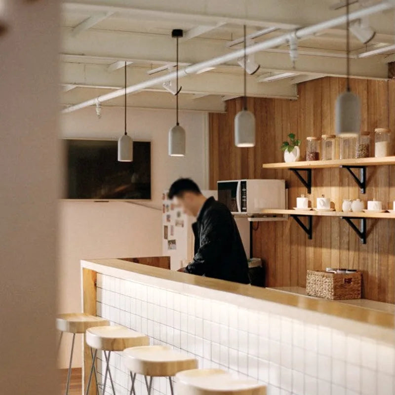 LUXELY cement pendant lights installed above a wooden bar counter in a cozy café. Industrial lighting for restaurant or hotel use.
