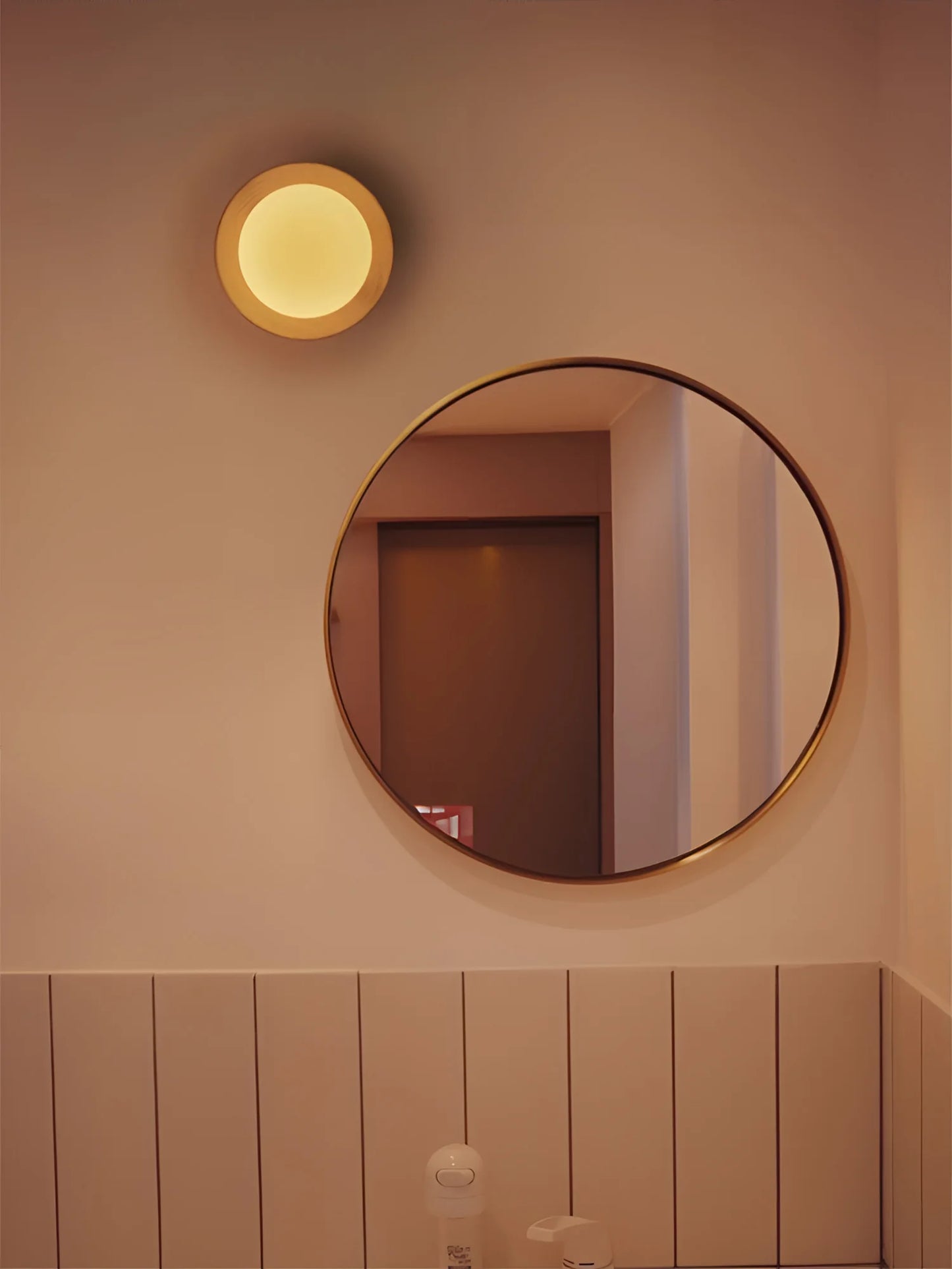 Light wood wall sconce above round mirror in bathroom, offering soft and stylish lighting for vanity areas.