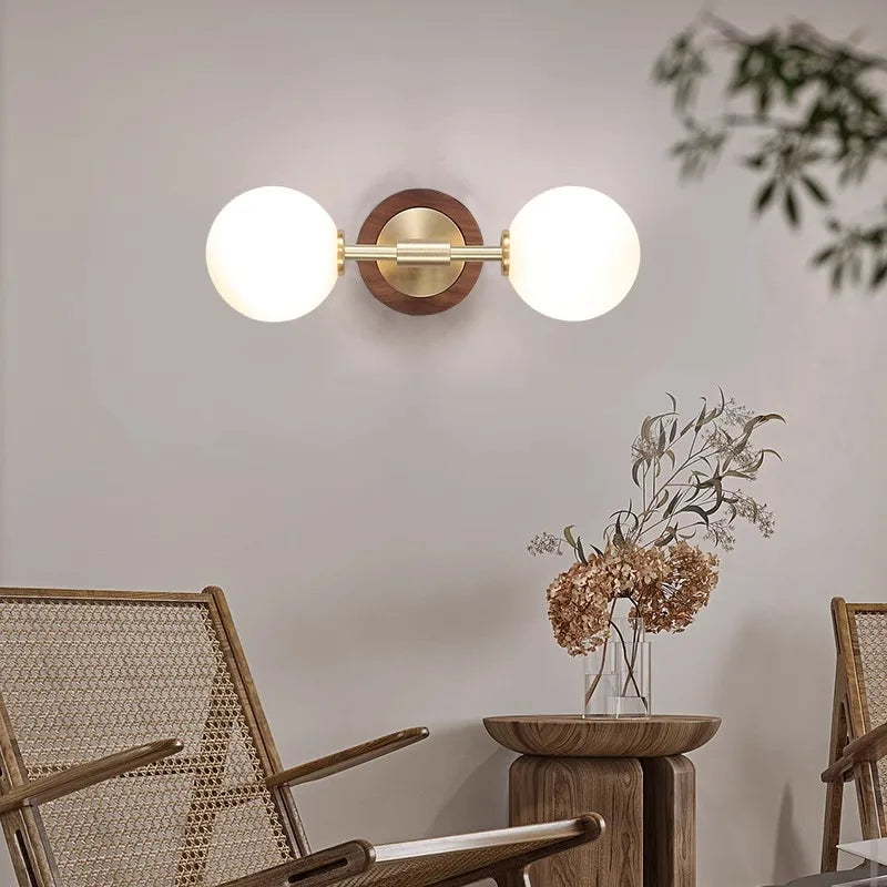Horizontal brass wall sconce with two frosted globes, styled in a cozy seating area with wooden furniture and dried flowers.