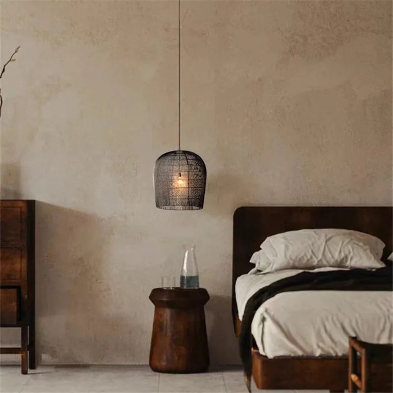 Single black LUXELY rattan pendant lamp beside a wooden bed, offering warm, ambient lighting in a rustic bedroom setting.