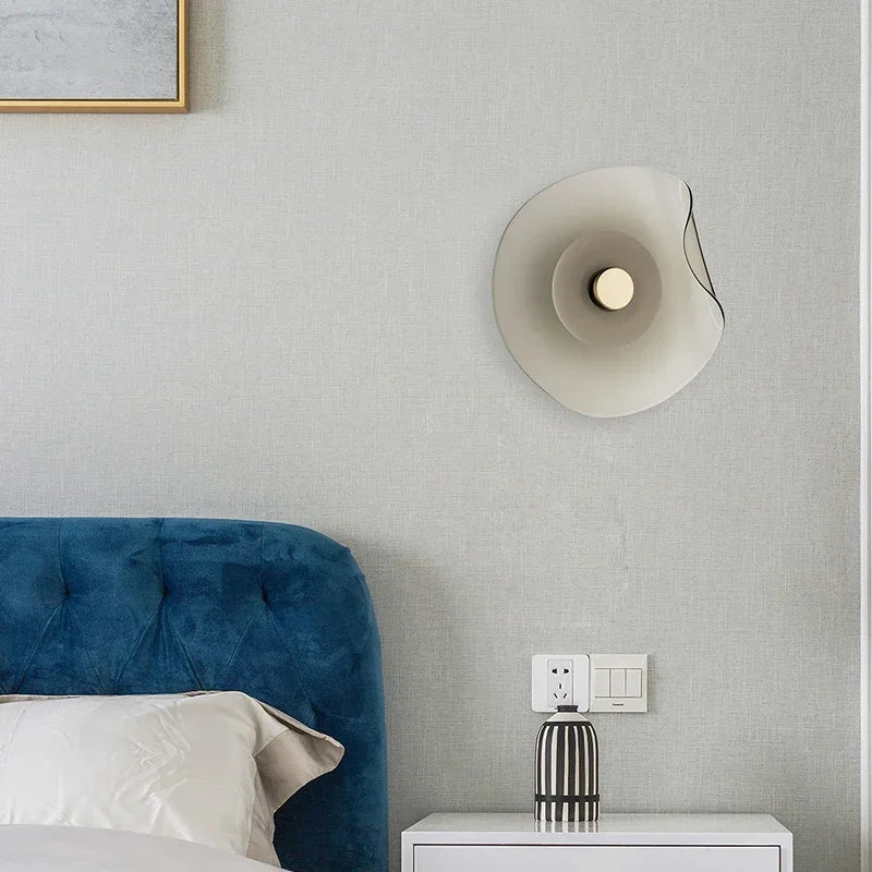 Modern glass wall lamp with round art deco design installed above a nightstand, enhancing bedroom ambiance with soft LED lighting.