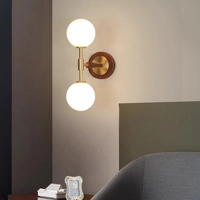 LUXELY brass wall lamp with vertical dual globe design, placed beside a modern bed for soft ambient bedroom lighting.