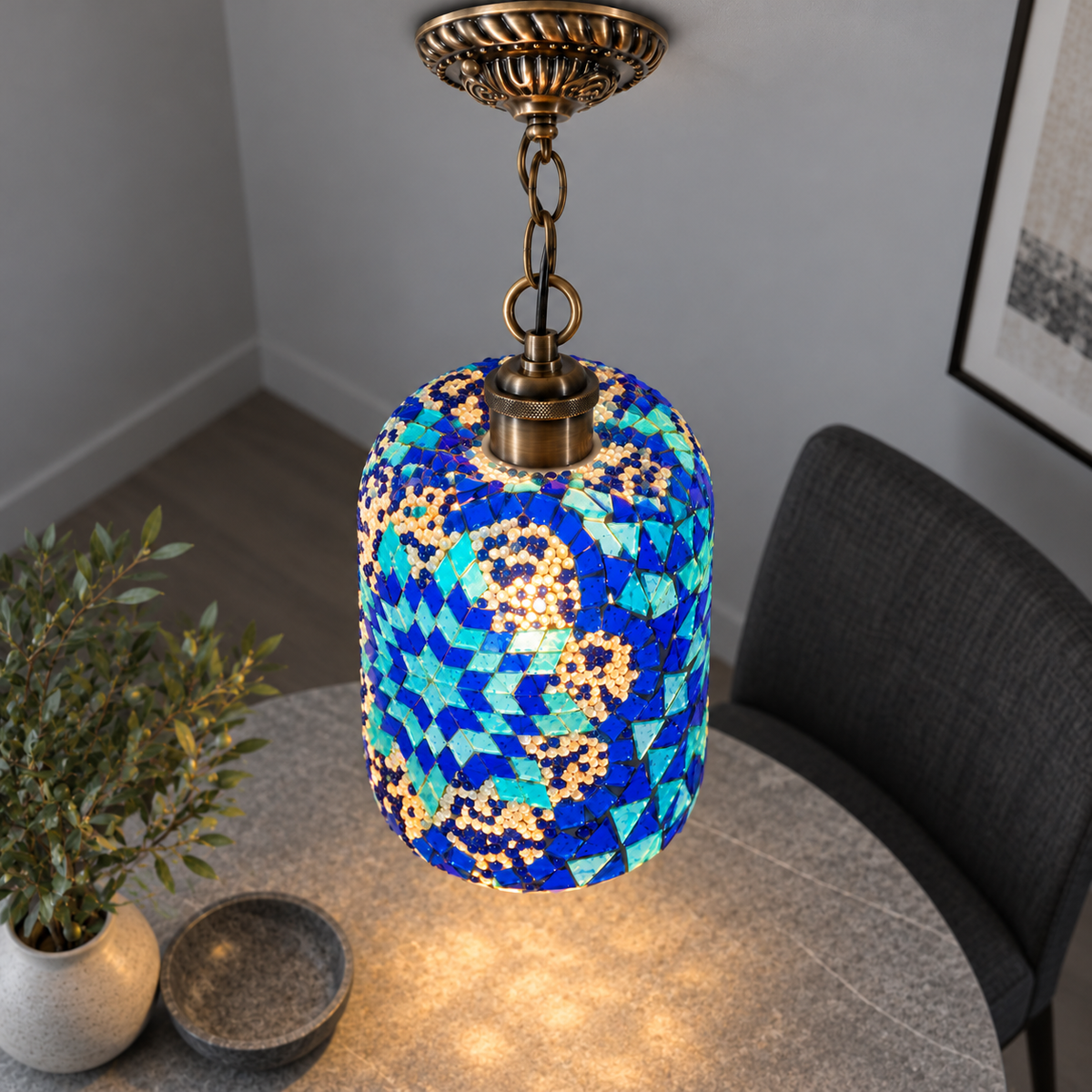 Illuminated Style C mosaic cylinder pendant with bronze chain, presented from an elevated side angle in a neutral grey room.