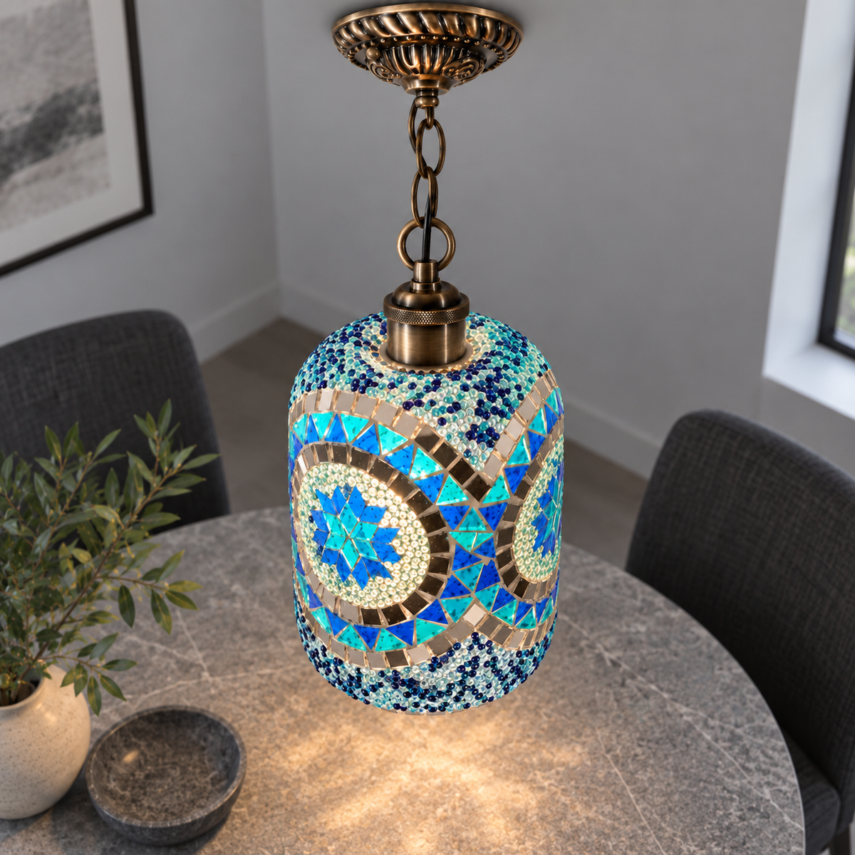 Illuminated Style H mosaic cylinder pendant with bronze chain, presented from an elevated side angle in a neutral grey room.