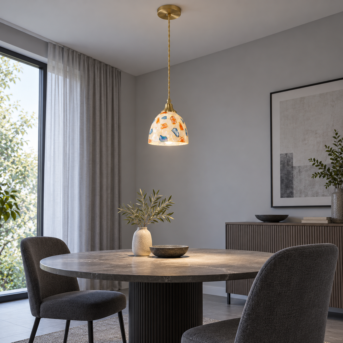 Small multicolour shell and glass dome pendant lit above a stone dining table, viewed from the side across a grey room.